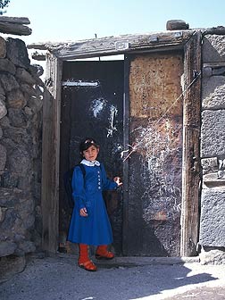 気の門扉の前に、青いワンピースの制服を着た小学生の女の子。真っ赤な靴がアクセントだ。